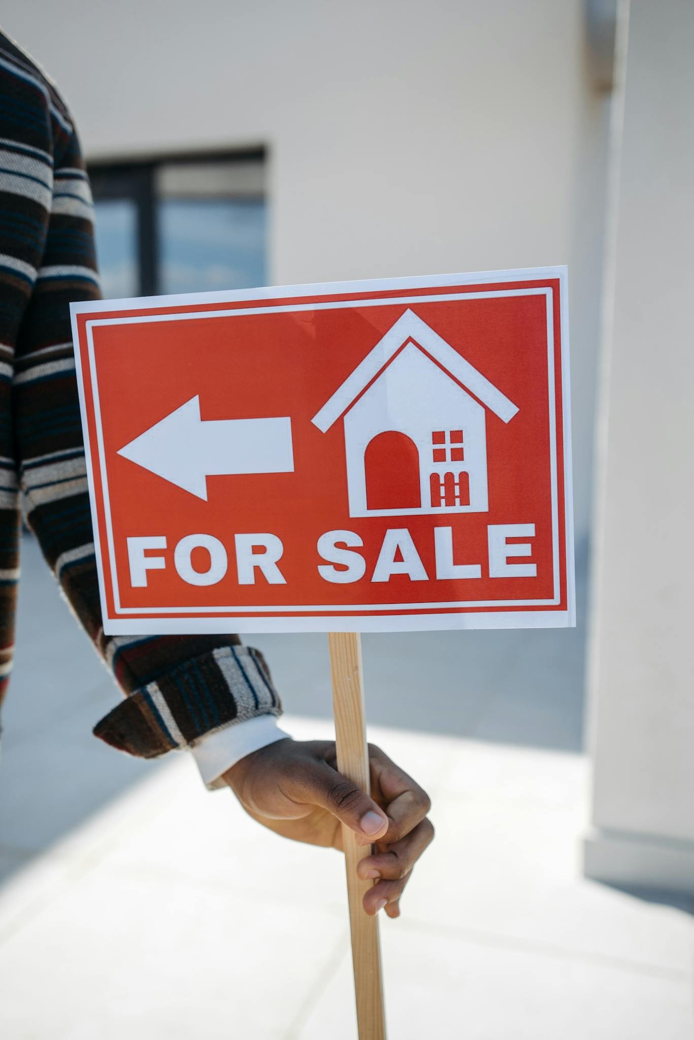 Close-up of a hand holding a 'For Sale' real estate sign with a house icon outdoors.